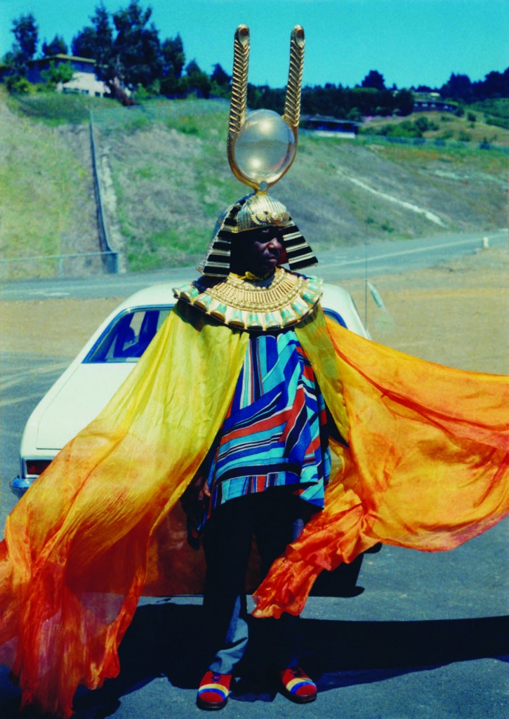 Faded photo of Sun Ra striding down a road in an orange cape, multicolored clothes, and a tall Egyptian-inspired headdress